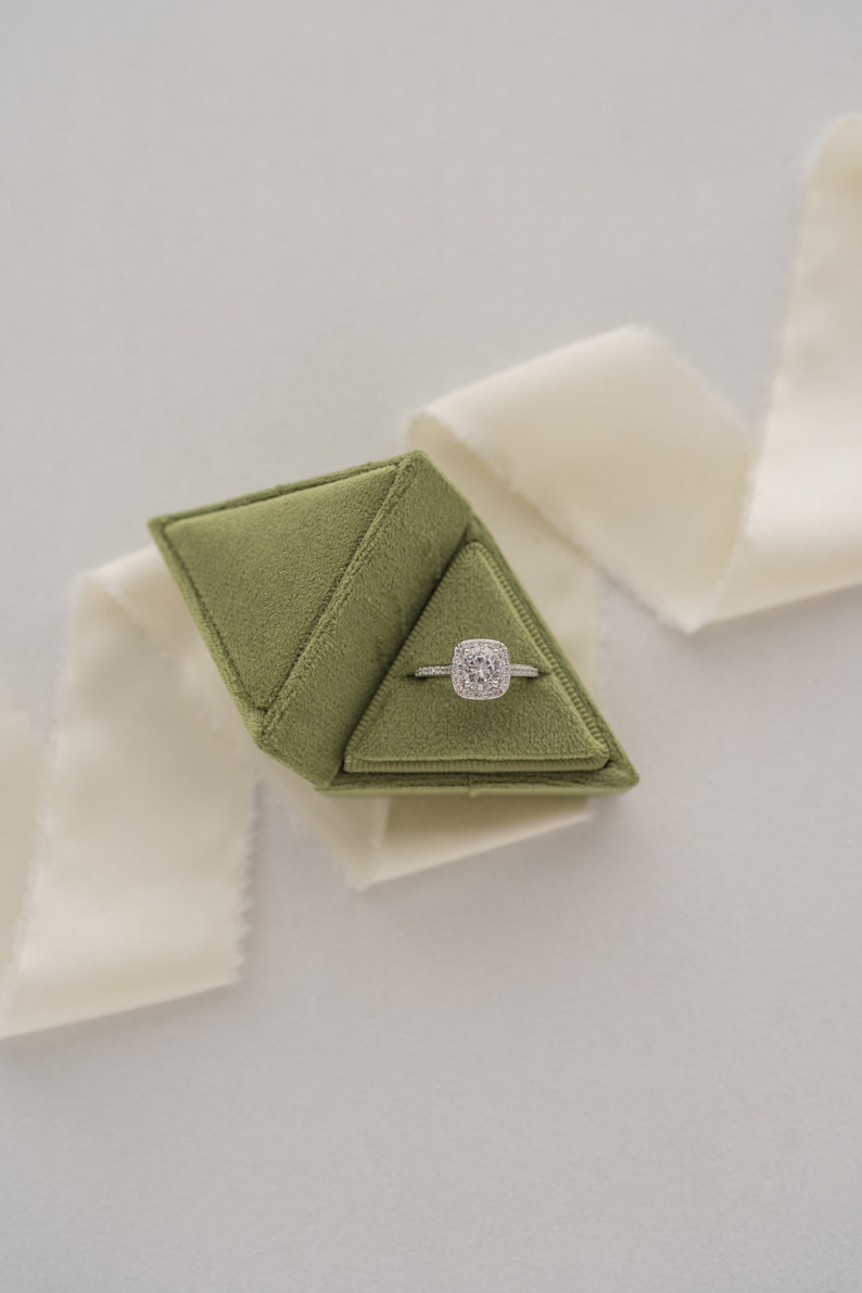 May include: A green velvet ring box with a diamond engagement ring inside. The box is shaped like a pyramid and is sitting on a white surface.