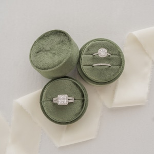 May include: Three green velvet ring boxes with a white satin ribbon. Two boxes contain engagement rings with a diamond center stone and a band of smaller diamonds. The third box is empty.