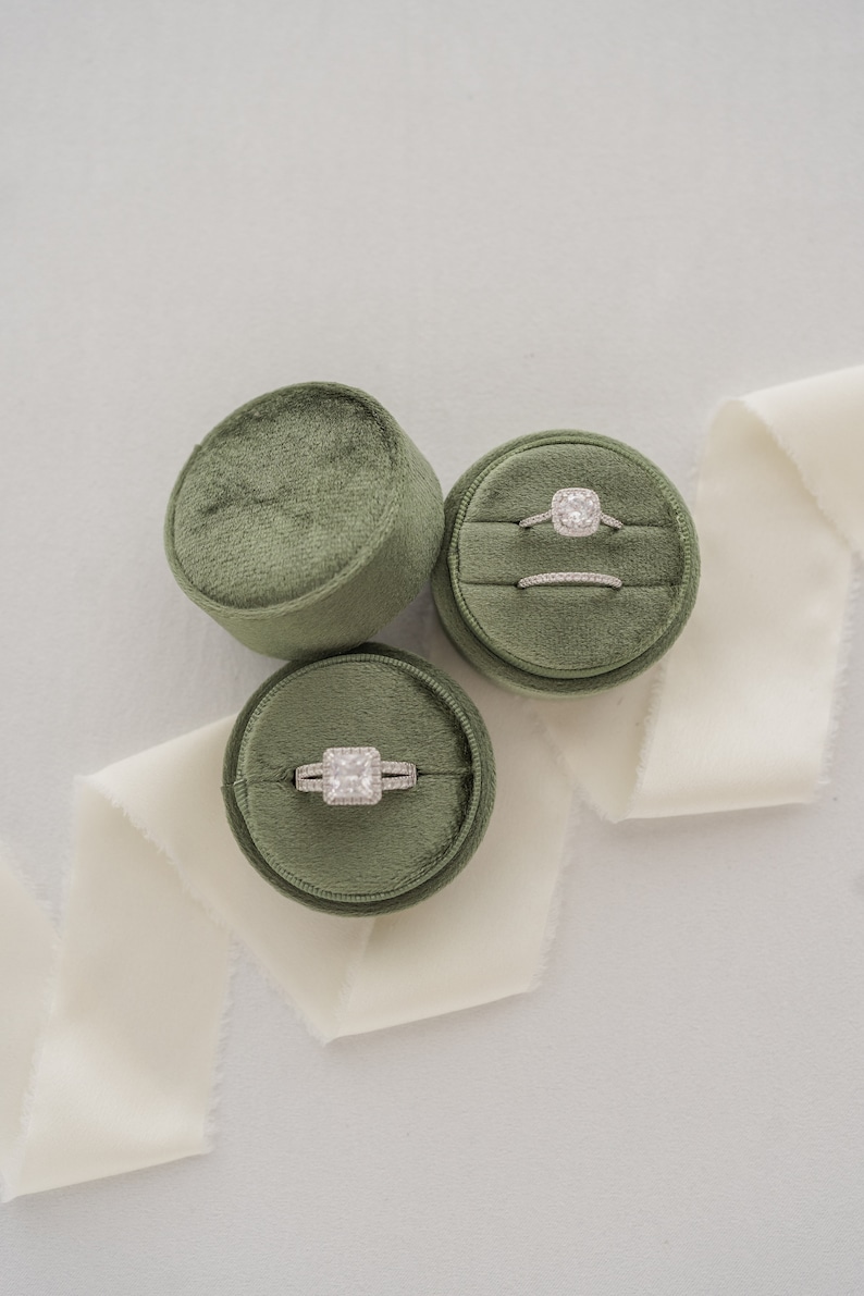 May include: Three green velvet ring boxes with a white satin ribbon. Two boxes contain engagement rings with a diamond center stone and a band of smaller diamonds. The third box is empty.