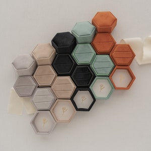 May include: A collection of hexagonal ring boxes in various colors, including black, beige, gray, and orange. Some boxes are open, revealing a floral design. The boxes are arranged on a white surface with a cream-colored ribbon.