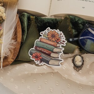 May include: A sticker of a stack of four books with floral accents. The books are labeled "Cozy Mystery", "Paranormal Romance", and "Dark Academia".