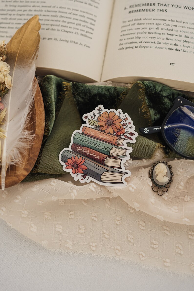 May include: A sticker of a stack of four books with floral accents. The books are labeled "Cozy Mystery", "Paranormal Romance", and "Dark Academia".