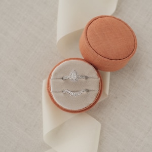 May include: Two rings in a small, round, orange velvet ring box. The top ring is a pear-shaped engagement ring with a diamond center stone and smaller diamonds around the band. The bottom ring is a delicate band with small diamonds set in a curved shape.