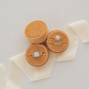 May include: Two velvet ring boxes in a mustard yellow color with a white satin ribbon. One box contains a square-shaped engagement ring with a halo setting, and the other box contains a wedding band.