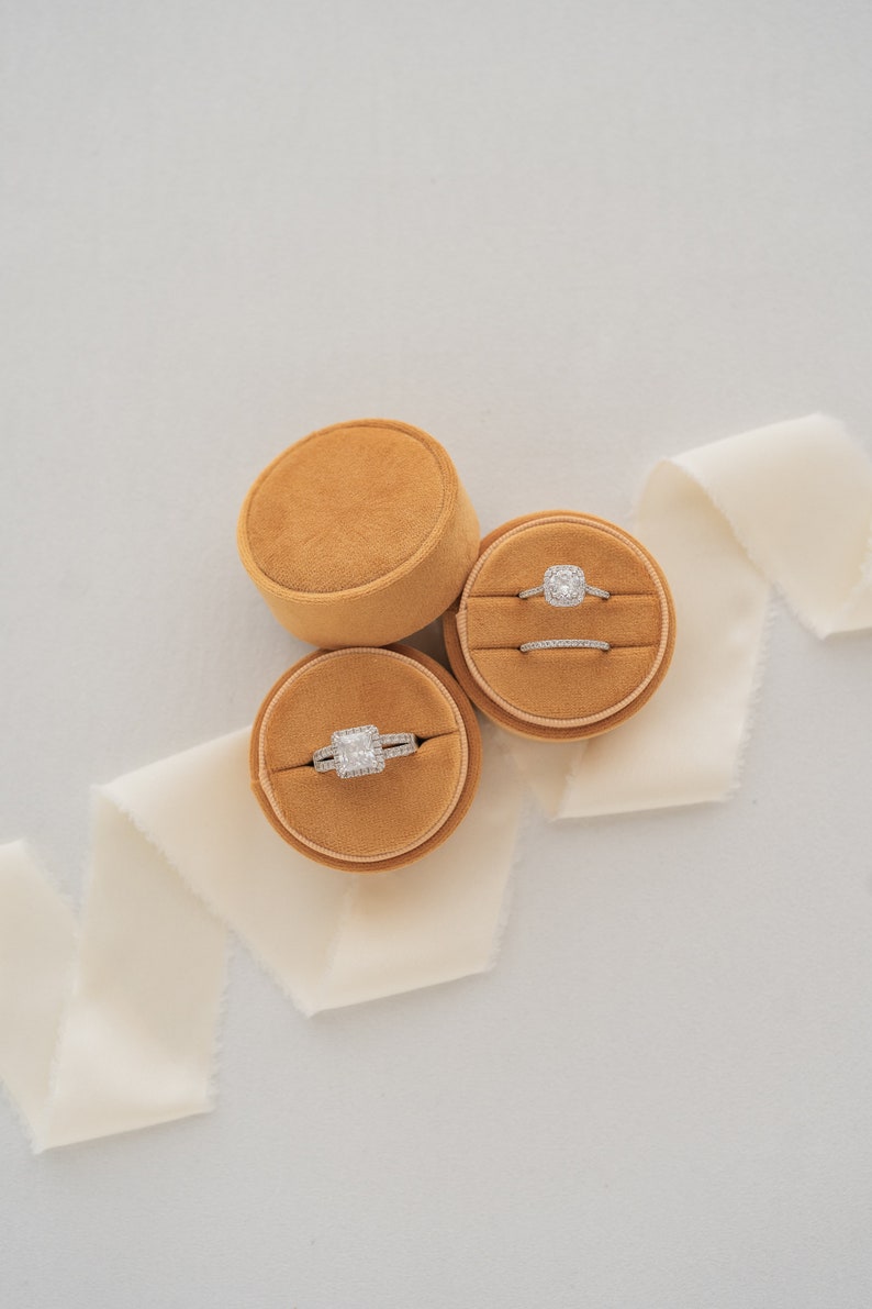 May include: Two velvet ring boxes in a mustard yellow color with a white satin ribbon. One box contains a square-shaped engagement ring with a halo setting, and the other box contains a wedding band.