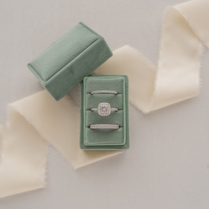 May include: A green velvet ring box with four rings inside. The top ring is a square-shaped engagement ring with a large center stone and smaller stones around it. The other three rings are simple bands with small stones.