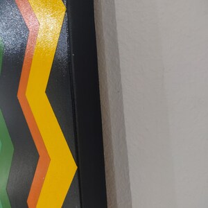 Może przedstawiać: Zbliżenie na kolorowy obraz abstrakcyjny w czarnej ramie. Obraz przedstawia wz&oacute;r w jodełkę w odcieniach zieleni, pomarańczy i ż&oacute;łci.