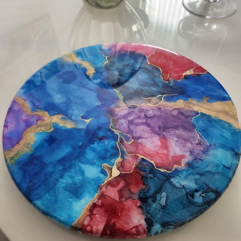 May include: A circular decorative tray with an abstract design. It features a mix of blue, purple, and red colours, highlighted by gold-coloured lines. The tray has a glossy finish, making it a striking decorative item.