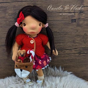 Könnte beinhalten: Handgefertigte Puppe mit schwarzen Zöpfen, trägt einen roten Pullover und einen gemusterten Rock. Die Puppe hat eine braune, fuchsähnliche Tasche mit einem Teddybär. Der Text "Aurélie & Flocke" ist sichtbar.
