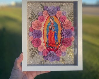 May include: A framed picture with a religious image of the Virgin Mary surrounded by paper flowers in shades of pink, purple, and white. The flowers are arranged in a circular pattern around the image.