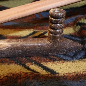 May include: A brown stone pipe with a cylindrical bowl and a stacked ring design on top. A wooden stick rests above the pipe. The pipe is set on a patterned, textured surface.