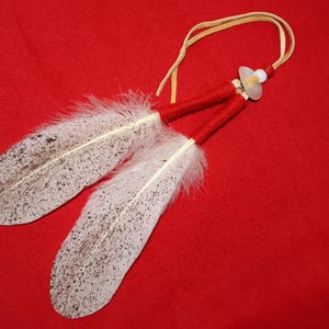 May include: Two white feathers with black speckles are tied together with red and white cord. The feathers are attached to a leather cord with a white bead.
