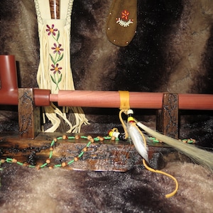 May include: A wooden pipe stand with a red and brown pipe resting on it. The stand is decorated with beads and a feather.
