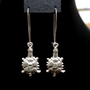 May include: A pair of silver-toned earrings. Each earring features a long, thin hook and a pendant shaped like a turtle. The turtle pendant has a detailed design with a textured shell and a small, circular emblem in the center.