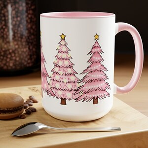May include: White ceramic mug with a pink rim and handle. The mug features a design of four pink Christmas trees with yellow stars and white lights.