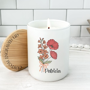 May include: A white cylindrical candle in a glass jar with a lit wick. The front features a floral illustration of red and orange flowers tied with a ribbon, and the name Patricia printed below. A round bamboo lid with the text CUSTOM ENGRAVED LID sits beside it.