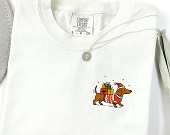 May include: An ivory-colored t-shirt with a Comfort Colors label at the neck. The shirt features a small graphic on the chest of a brown dachshund wearing a red and white patterned sweater and a Santa hat, carrying stacked colorful gift boxes.