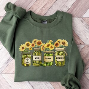 Peut inclure: Un sweat-shirt vert militaire avec une impression graphique de quatre bocaux en verre remplis de cornichons verts, entremêlés de tournesols jaunes. Chaque bocal porte une étiquette avec le texte : JUST A GIRL, WHO, LOVES et PICKLES.