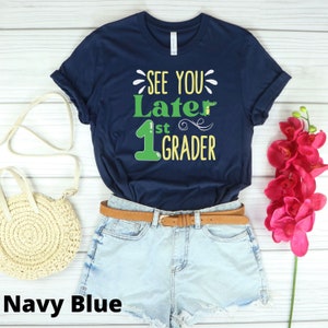 May include: A navy blue short-sleeve t-shirt featuring the text "SEE YOU Later 1st GRADER" in white, green, and yellow, accompanied by a small green alligator graphic. The shirt is paired with light blue denim shorts and a brown belt.