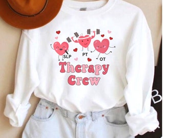 May include: A white long-sleeve sweatshirt featuring a graphic of three cartoon hearts with arms and legs. The hearts are labelled SLP, PT, and OT. Below the hearts, the text reads Therapy Crew in pink and red letters, surrounded by small hearts.
