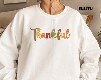 Puede incluir: Una sudadera blanca de cuello redondo con la palabra Thankful impresa en el pecho en una fuente cursiva. Cada letra de la palabra está coloreada en varios tonos de naranja, amarillo, marrón y verde, creando un diseño inspirado en el otoño.