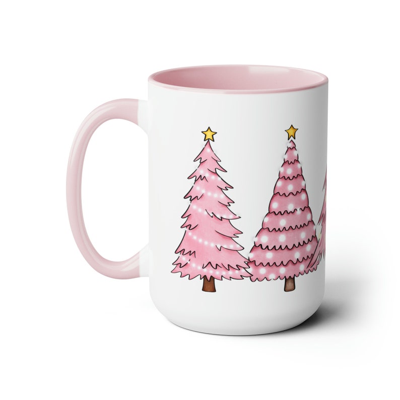 May include: A white ceramic mug with a pink interior and handle. The mug features a design of three pink Christmas trees with white dots and yellow stars on top.