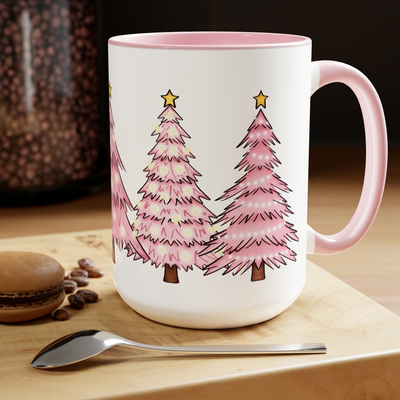 May include: White ceramic mug with a pink rim and handle. The mug features a design of four pink Christmas trees with yellow stars and white lights.