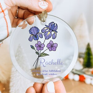 Puede incluir: Un adorno de cristal transparente con un dise&ntilde;o floral con iris moradas y flores moradas m&aacute;s peque&ntilde;as. El nombre "Rachelle" est&aacute; escrito en una fuente de escritura debajo de las flores, con palabras como "modestia" y "virtud". El adorno tiene una cinta dorada.