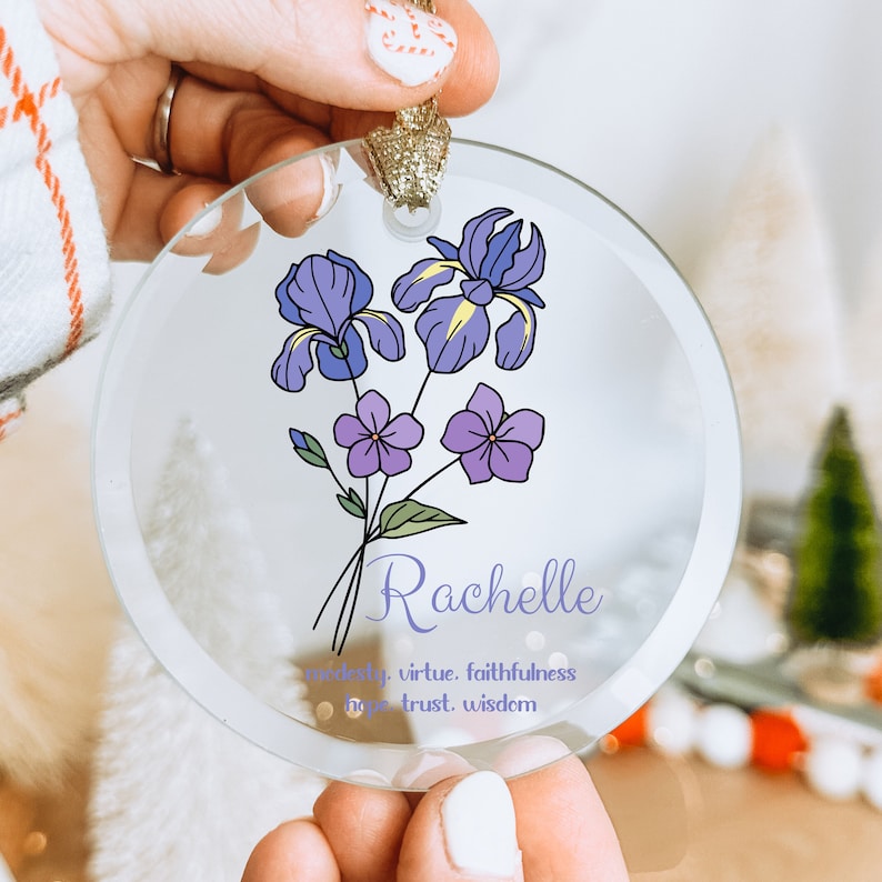 Puede incluir: Un adorno de cristal transparente con un dise&ntilde;o floral con iris moradas y flores moradas m&aacute;s peque&ntilde;as. El nombre "Rachelle" est&aacute; escrito en una fuente de escritura debajo de las flores, con palabras como "modestia" y "virtud". El adorno tiene una cinta dorada.