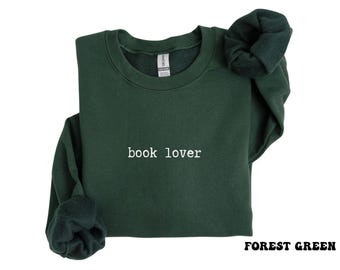 Puede incluir: Una sudadera de cuello redondo en color verde bosque con el texto "book lover" bordado en hilo blanco en el centro del pecho. La prenda tiene mangas largas con puños acanalados y un cuello acanalado, mostrando un forro interior de felpa suave.
