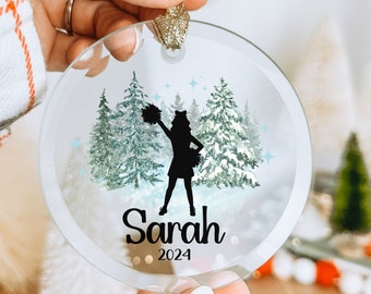 May include: A clear circular glass ornament featuring a black silhouette of a cheerleader holding pom-poms, set against a watercolor forest of pine trees with light blue star accents. The name Sarah and the year 2024 are printed in black text.