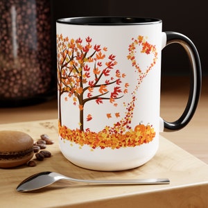 May include: White ceramic mug with a black interior and handle. The mug features a fall-themed design with a tree, falling leaves, and pumpkins in shades of orange, red, and yellow. A spoon and a pastry are in the foreground.