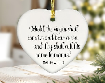 May include: A white heart-shaped ceramic ornament featuring black printed text that reads, "Behold, the virgin shall conceive and bear a son, and they shall call his name Immanuel" followed by "MATTHEW 1:23". It hangs from a gold-colored string.