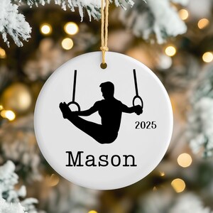 May include: A round white ceramic Christmas ornament featuring a black silhouette of a gymnast performing on rings. The text "Mason" is printed in black at the bottom, and the year "2025" is printed in black to the right of the gymnast figure.