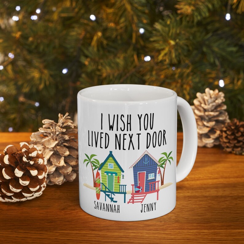 Puede incluir: Taza de cer&aacute;mica blanca con un gr&aacute;fico en blanco y negro de dos casas de playa con palmeras y el texto "Ojal&aacute; vivieras al lado". Las casas est&aacute;n etiquetadas como "Savannah" y "Jenny".
