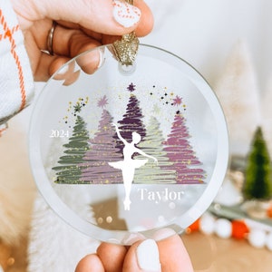 May include: A clear glass Christmas ornament with a white silhouette of a ballerina dancing in front of a pink, green, and gold glitter tree design. The ornament also includes the text "2024" and "Taylor".