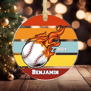 May include: A round Christmas ornament featuring a baseball engulfed in flames, with the text "2024" and the name "BENJAMIN". The background has horizontal stripes of orange, yellow, and blue.