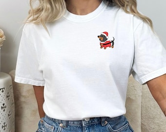 May include: A white short-sleeved t-shirt featuring a small graphic on the left chest area. The graphic depicts a brown and black dachshund dog wearing a red Santa hat and a red coat with a black belt and white trim, standing in profile.