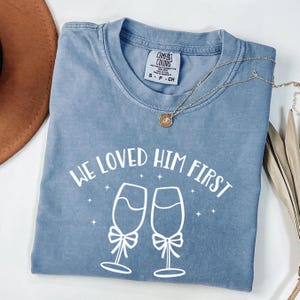May include: A blue short-sleeved t-shirt with a white graphic print featuring two champagne flutes with bows and the text "WE LOVED HIM FIRST" with small star accents. The shirt has a Comfort Colors brand label at the inner neckline.