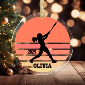 May include: A round, clear glass ornament with a silhouette of a softball player against a sunset background. The ornament has the year "2024" and the name "Olivia" printed on it.