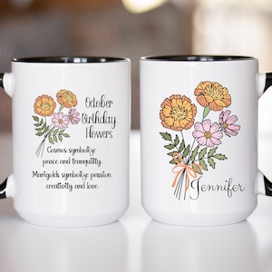 May include: A white ceramic mug with a black interior and handle, featuring a floral illustration of orange marigolds and pink cosmos. The text reads October Birthday Flowers, Cosmos symbolize peace and tranquility, Marigolds symbolize passion, creativity and love, and the name Jennifer.