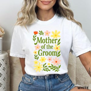 May include: A white short-sleeve t-shirt featuring a central graphic with the green text Mother of the Groom surrounded by various colourful flowers in shades of yellow, orange, and pink, along with green leaves and stems on the front.