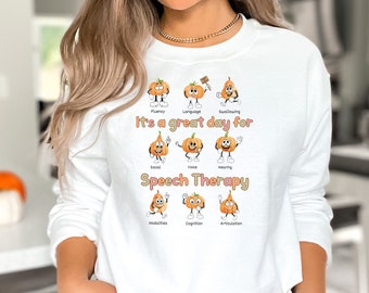 May include: A white long-sleeve sweatshirt featuring a graphic of nine cartoon pumpkins with arms and legs. The text reads "It's a great day for Speech Therapy" with labels for Fluency, Language, Swallowing, Social, Voice, Hearing, Modalities, Cognition, and Articulation.