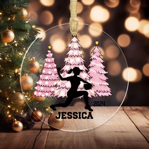 May include: Clear acrylic Christmas ornament featuring a black silhouette of a softball player in action, with pink Christmas trees and the name "JESSICA" below. The year "2024" is also printed on the ornament. A gold ribbon is attached for hanging.