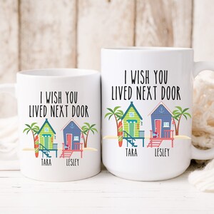 Puede incluir: Dos tazas de caf&eacute; blancas con el texto "Ojal&aacute; vivieras al lado" y una ilustraci&oacute;n de una casa de playa. Las tazas tienen los nombres "Tara" y "Lesley" escritos en las casas de playa.