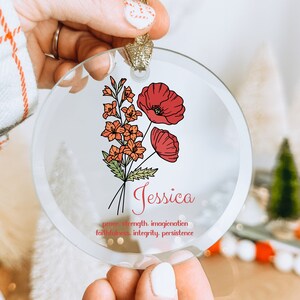 Puede incluir: Adorno navide&ntilde;o redondo de cristal con un dise&ntilde;o floral. Presenta amapolas rojas y flores naranjas, con el nombre "Jessica" en escritura roja. Tiene una cinta dorada para colgar.