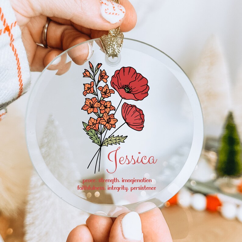 Puede incluir: Adorno navide&ntilde;o redondo de cristal con un dise&ntilde;o floral. Presenta amapolas rojas y flores naranjas, con el nombre "Jessica" en escritura roja. Tiene una cinta dorada para colgar.