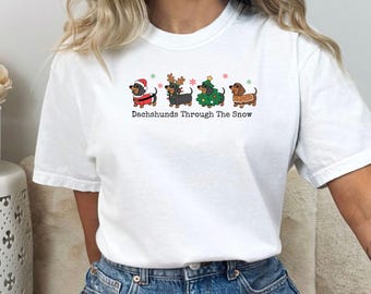 May include: A white t-shirt featuring a graphic of four dachshunds in holiday attire, including a Santa suit, reindeer antlers, a Christmas tree costume, and a gingerbread sweater, with the text "Dachshunds Through The Snow" printed below.