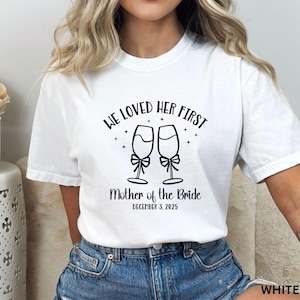 May include: A white t-shirt featuring a black graphic of two champagne flutes with bows. The text reads "WE LOVED HER FIRST", "Mother of the Bride", and "DECEMBER 3, 2025". The shirt is paired with blue denim jeans, showcasing a casual and clean aesthetic.