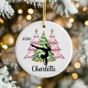 May include: A round white ceramic ornament featuring a black silhouette of a gymnast performing a handstand. The design includes two pink Christmas trees and one green Christmas tree, the year 2025, and the name Charlotte in black script.
